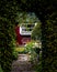 View through a an arch of ivy into a mystic garden with flowers, a bench and a red facade with white window in summer time