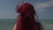 View of the back of a woman with red hair flying in the wind against the backdrop of the sea surf