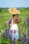 View from the back of a young girl in a dress and hat with a bouquet of lupins in her hands on a background of green meadow