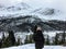 A view from behind of a female hiker alone on top of a mountain admiring a beautiful snow covered mountain and forest