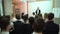 View from behind of a group of students in a classroom, listening as their teacher holds a lecture. One student puts up