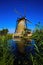 View beyond green reed grass over water canal on one isolated old windmill with reflection against deep blue cloudless summer sky