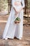 View of bride in white attire holding wedding bouquet in forest