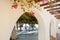 A view of bright white building with arch and pink violet bougainvillea and green trees in the street of small mediterranean town