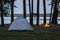 View of the campsite by the lake at twilight. Tent and burning fire place by the water surrounded by trees in the forest. Spending