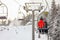 View from chairlift over ski piste, skier in bright red jacket seating in front, more blurred people skiing below, active snowguns