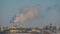 View on city buildings and smoke from factory smokestack against blue sky