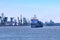 View of commercial cargo port with container bridge cranes and a ship entering port on a sunny day.