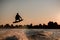 view of dark silhouette of active male rider holds rope and making extreme jump on wakeboard at sunset.