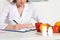 View dietitian in white coat writing in clipboard at workplace with pills, fruits and vegetables on table