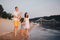 View Of Family With Two Toddler Children Outdoors By The River In Summer. Happy Young Family Have Fun On Beach At Sunset
