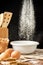 View of flour falling on white bowl with rustic wooden objects, on wooden table, on black background