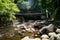 View of fresh river stream flow, stone bank and natural rock cascade with light glare around green trees and bridge in Kurokawa