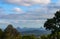 View of the Glasshouse Mountains in Queensland Australia framed by trees - under cloudy blue skies with a fire in the valley