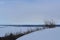 View from high snowy bank on frozen river and other bank  through leafless trees