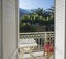 View from hotel room balcony door over white table and red bamboo chair and flowering bush and palm trees on background