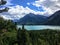 A view of a majestic turquoise lake surrounded by vast green evergreen forests and mountains on a sunny day with blue sky.