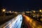 View of multilane urban highway with glowing lines of light from car headlamps and modern architecture