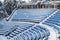 View of outdoor amphitheater rows of blue plastic seats covered in snow at winter time on sunny day