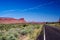 View over green valley on endless straight road with steep red sandstone hill formation butte against blue sky