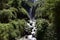 View of Peguche Waterfall in the mountains. It's surrounded by green forest full of vegetation. Ecuador