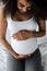 View of positive curly african american pregnant woman touching belly while sitting on bed