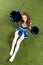 View of pretty cheerleader girl in blue uniform lying with pompoms on green field