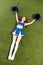 View of pretty cheerleader girl in blue uniform lying with pompoms on green field