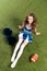View of pretty cheerleader girl in blue uniform lying with pompoms and rugby ball on green field