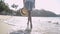 View from rear side of asian girl holding straw hat in casual dress with barefoot walking on the beach under sunlight.