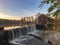 View of the reflecting pond, waterfall, wooden building and water wheel at sunset in autumn under an orange and blue sky at