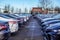 View of rows of various colored new cars in a parking lot with a access space between them.