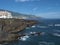 View on sea shore with lava rock cliffs at village Los Cancajos, ocean and green hills. Blue sky background. La Palma