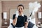 View of smiling pretty maid in white gloves holding duster and talking on smartphone in hotel room