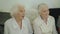 View of two grey-haired, cute senior women in white blouses talking in room