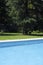 View of various materials by a pool. Blue and dark blue water, a concrete edge, paving slabs, green lawn. A tree in the