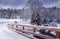 View of white winter wonderland scenery in Vermont with snow covered trees, bench, pond, small house, road and river.