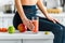 View of woman holding glass of smoothie while sitting in kitchen near apples