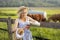 Village girl pouring milk into a glass, on the background of fields with grazing cows. Summer rural life in Germany.