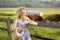 Village girl pouring milk into a glass, on the background of fields with grazing cows. Summer rural life in Germany.