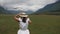 village woman in a white dress and hat walks through a green field among the mountains in summer.