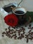 Vintage beautiful cup of hot coffee, bright red rose, green leaves, scattered coffee beans and dark chocolate bar on a wooden tabl