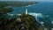 Vintage Dutch lighthouse stands on tropical island with greenery. Aerial drone big ocean waves hitting rocky coast