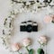 Vintage film camera in the middle, sakura branch, pink rose flowers on the white wooden desk. Top view, flat lay