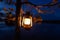 Vintage oil lamp hanging on a tree. Beautiful view of dark forest and lake at night. Hiker, Travel, Outdoor Concept