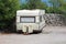 Vintage old dilapidated camper trailer parked in backyard on gravel surface next to improvised stone wall surrounded with dense