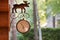 Vintage style street clock with metal deer sign hanged on a wooden log wall with green park or backyard on the background