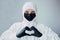 Virologist in a black mask, medical gloves and dressed in viral protective  suit shows a heart with hands on a gray background. Is