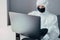 A virologist in a black mask and medical gloves dressed in a white protective suit works with a laptop and looks for information a