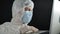 Virologist in a medical mask, rubber gloves and a protective suit works at a laptop on a dark background. Development of